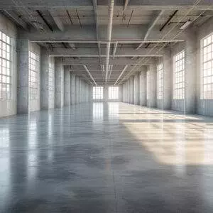 A large empty industrial warehouse in Oregon features a polished concrete floor, enhanced by a durable concrete coating. With large windows on both sides and visible ceiling pipes and beams, sunlight streams in, creating a striking pattern of light and shadows.