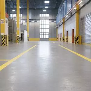 The interior of this empty industrial warehouse in Oregon showcases a polished concrete floor, marked with yellow lines. The space features high ceilings, large windows, and multiple loading bay doors, all perfectly enhanced by durable concrete coating for long-lasting quality.